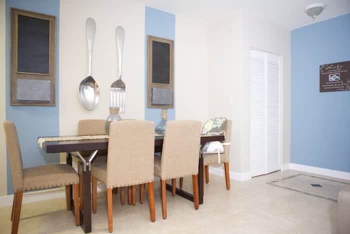 The dining room, set for six with beige upholstered chairs, showcases a harmonious blend of blue and white walls. Adorning the space are oversized spoon and fork decor units, while a sideboard with ornaments and a decorative frame complete the inviting ambiance.