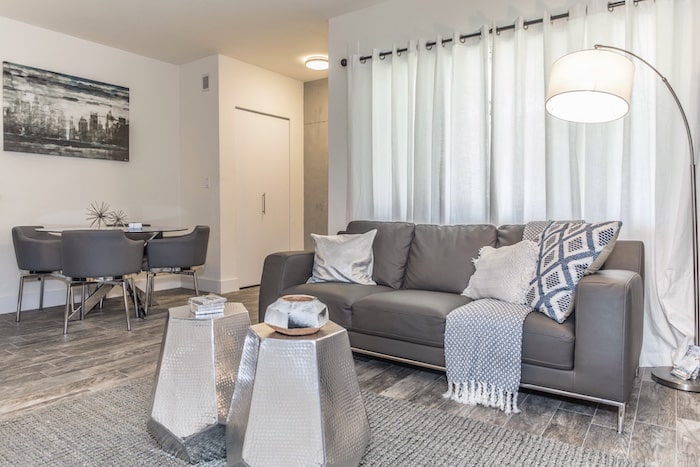 A modern living room with a gray sofa adorned with pillows and a blanket. Two metallic hexagonal coffee tables stand on a rug. A floor lamp and white curtains add light, while a dining area with a round table and chairs is visible in the background.