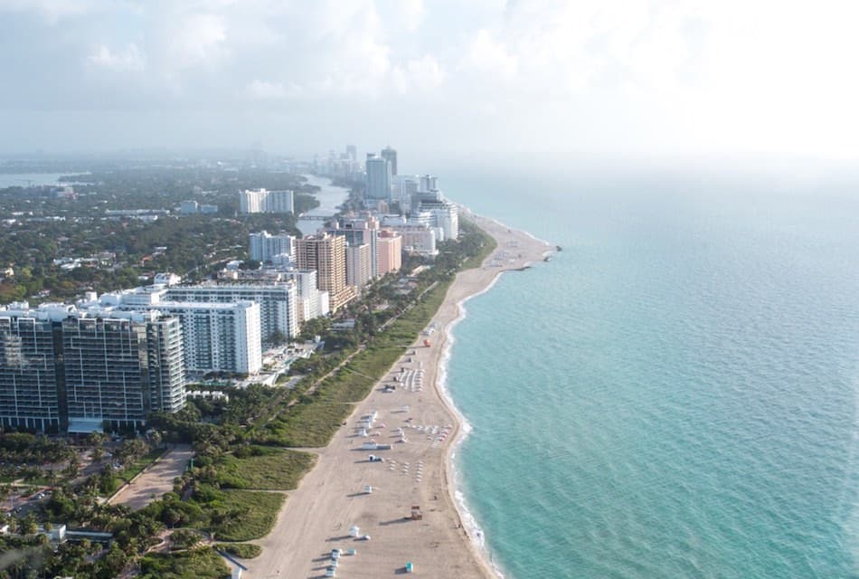 Aerial view of a coastal city with tall buildings lining a sandy beach. Sun umbrellas are scattered along the shoreline, and the turquoise ocean stretches towards the horizon under a partly cloudy sky.