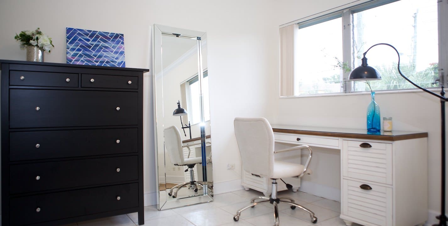 A modern, minimalist home office with a white desk and swivel chair near a large window. A tall black dresser with silver knobs stands to the left, and a large mirror leans against the wall. A floor lamp and blue artwork add decorative touches.