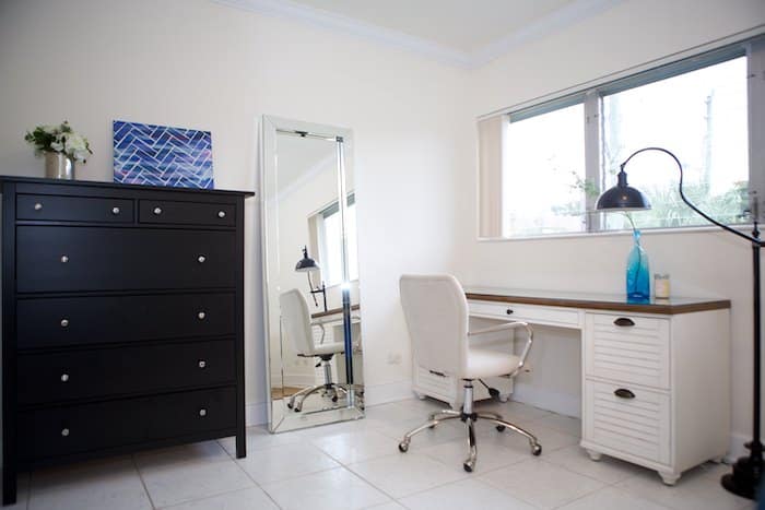 A tidy home office features a white desk and swivel chair, complemented by a black desk lamp. A blue decorative piece graces the desk. There's a large mirror, black storage units as drawers, and a potted plant. Natural light streams through the window.