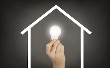 A hand holding a glowing light bulb is positioned inside a white outline of a house on a dark background, symbolizing innovation and ideas within the home.