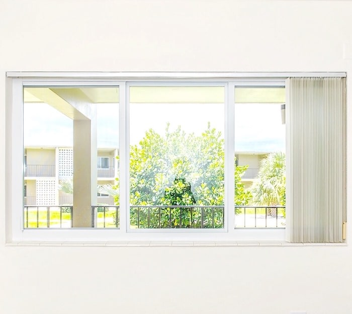 View through a large window with white frames, showing green trees and parts of a building outside. Vertical blinds are partially open on the right side. Bright, sunny day with a clear sky.