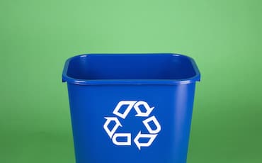 A blue recycling bin with a white recycling symbol on the front, set against a plain green background.