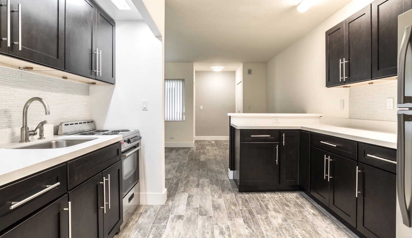 A modern kitchen with dark wood cabinets, white countertops, and stainless steel appliances, including a stove and refrigerator. The floor features gray tiles, and there's a view into a narrow hallway with natural light from a window.