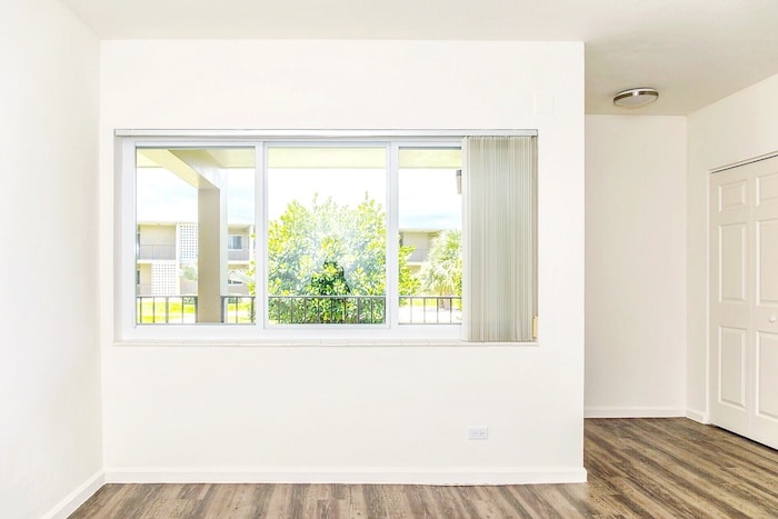 A bright, empty unit with white walls and hardwood flooring. A large window with vertical blinds offers a view of the green landscape outside. A closed white door is visible on the right, and a ceiling light fixture adorns the upper right wall.