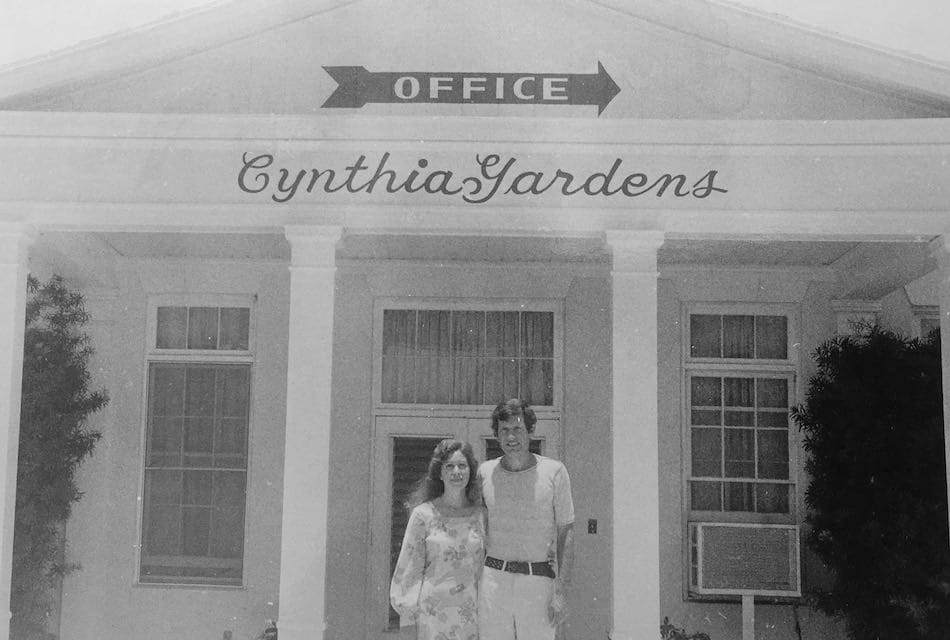 A black and white photo of a man and woman standing in front of a building with columns. The sign above reads "Office" with "Cynthia Gardens" below it. The building has large windows and an air conditioning unit visible on the right.