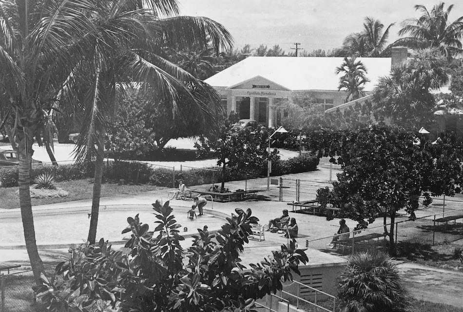 A black and white photo of a tropical scene with palm trees and bushes in the foreground. Several people are sitting and walking near a pathway. In the background, there is a building with a sign and more trees.