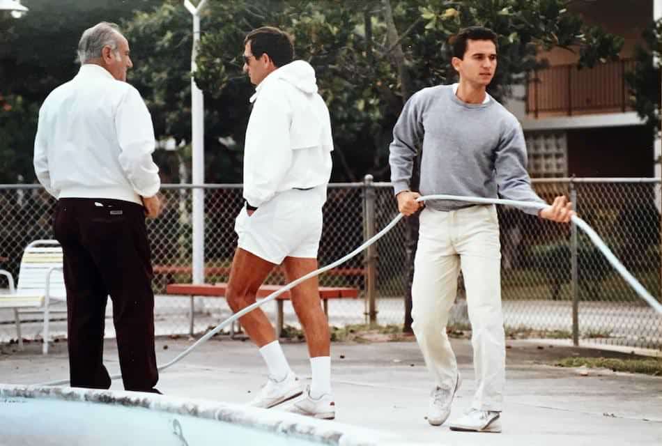 Three men are gathered poolside. One man in a grey sweater adjusts a hose, while the others in white athletic attire stand nearby, conversing. They appear to be near a chain-link fence with trees and buildings in the background.