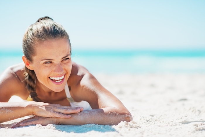 A woman with her hair tied back is lying on a sandy beach, smiling widely under the bright sun. The clear blue sea and sky are in the background, creating a serene and cheerful atmosphere.