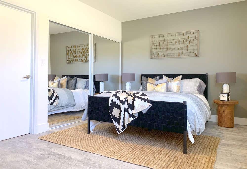 The modern bedroom features a woven black bedframe, gray bedding, and geometric pillows. A patterned blanket is draped on the bed. Two wooden side tables with lamps flank the bed, acting as sleek units of style. A large mirror and wall art enhance the decor while a jute rug covers the floor.