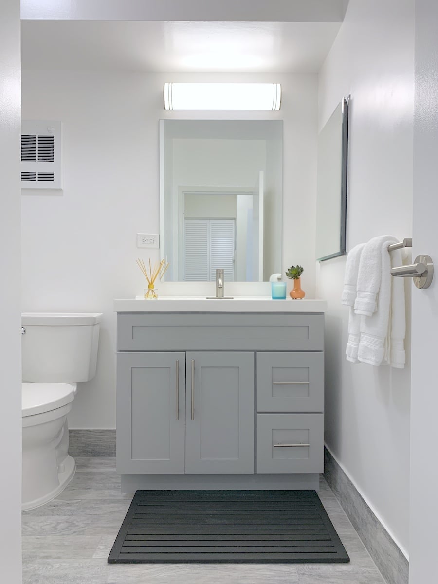 A modern, bright apartment bathroom features a gray vanity with a mirror above. It boasts a white countertop and sink, adorned with folded towels. A toilet sits to the left, while a small plant and diffuser add charm to the space.