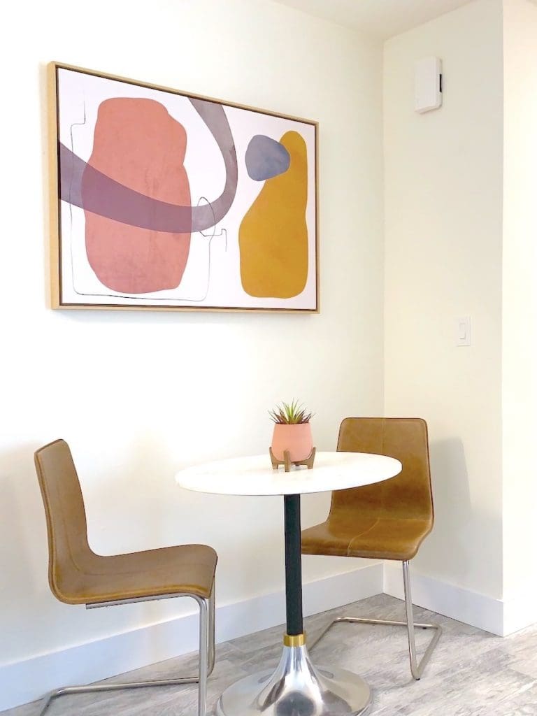 A small, modern dining area in a cozy apartment features a round white table with two brown chairs. A minimalist abstract painting in earthy tones adorns the wall above. Perfect for rental properties, a small potted plant adds charm at the table's center.