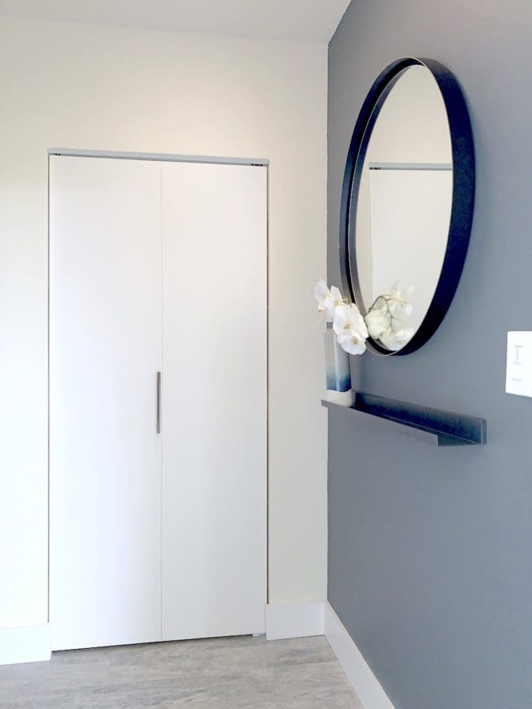 A minimalist apartment room with a gray accent wall features a round mirror and a small shelf holding white flowers. There's a white closet with simple handles on the adjacent wall and a light wooden floor.