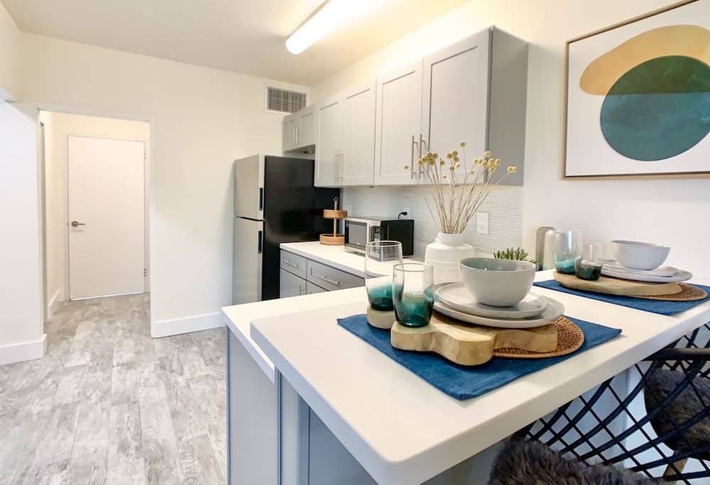 This modern apartment kitchen features sleek gray cabinets and a black refrigerator. A white counter is adorned with blue placemats, dishes, and glasses. A decorative plant and abstract artwork add a stylish touch, while the floor boasts a light gray wood finish.