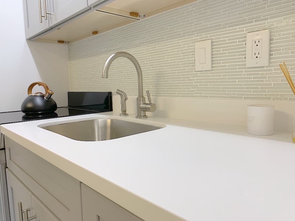 In contemporary apartments, a modern kitchen counter features a stainless steel sink, sleek faucet, and light gray mosaic tile backsplash. A black kettle sits on the stovetop beside a white candle. Pristine white cabinets with silver handles line the walls above and below.