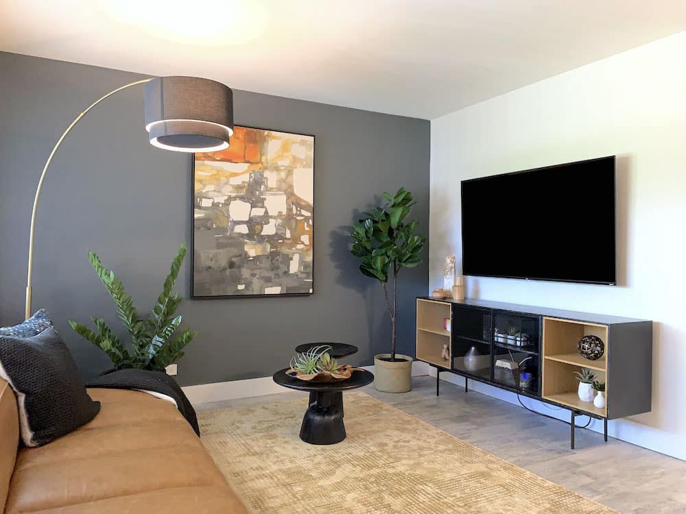 In a chic boca apartment, the modern living room features a brown leather sofa, abstract artwork on a dark accent wall, and a flat-screen TV mounted above a stylish console. A floor lamp arches over the seating area, while plants bring greenery to the contemporary space.