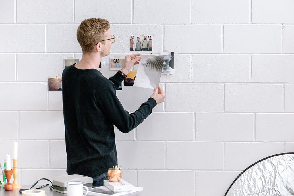 A person with glasses and a black shirt is standing against a white brick wall, pinning photos to it. The wall is adorned with various pictures. A table nearby holds books, a candle, and a decorative object.