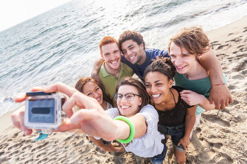 A group of six friends stands close together taking a selfie at a sunny beach location. They are smiling and casually dressed, with the ocean and sandy shore in the background. The clear sky enhances the cheerful atmosphere, highlighting the natural amenities of their seaside adventure.