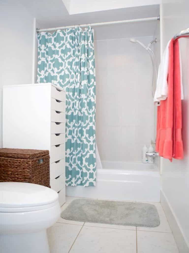 A bathroom reminiscent of a Martinique getaway, featuring a white toilet, wicker basket, and elegant white storage unit. The shower boasts a blue and white patterned curtain with towels neatly on the rail, complemented by a gray bath mat resting on the pristine tiled floor.