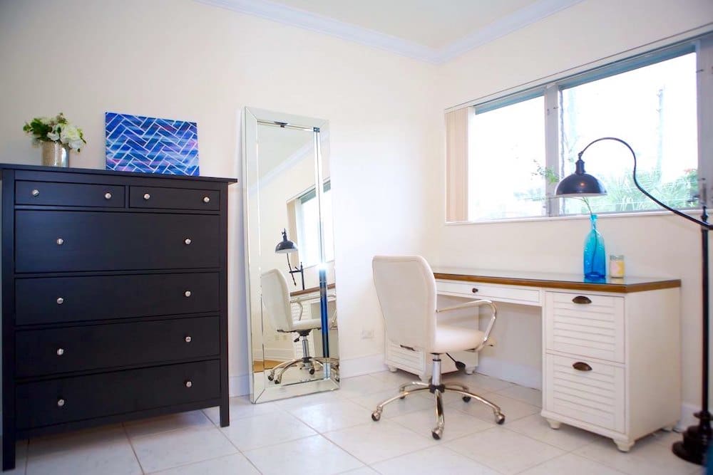 A minimalist home office with a white desk and chair, a black dresser, and a tall mirror exudes an airy Martinique charm. A blue vase and desk lamp sit atop the desk, while a painting and small plant adorn the dresser. Natural light streams through the window, enhancing the serene ambiance.