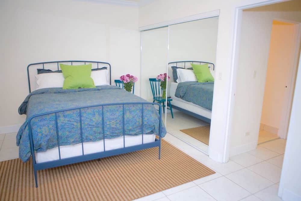 A bedroom with a blue metal bedframe, green and white pillows, and a blue floral comforter radiates a touch of Martinique elegance. A striped rug adorns the white tiled floor, while a large mirror reflects a chair with a vase of pink flowers. The door to the right leads to another room.