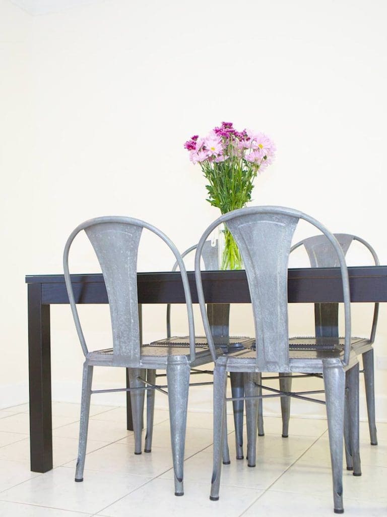 A modern dining room setup with a dark wooden table and three metal chairs evokes the elegance of Martinique. A vase with pink and purple flowers graces the table, while the background wall in a light cream color provides a serene backdrop.