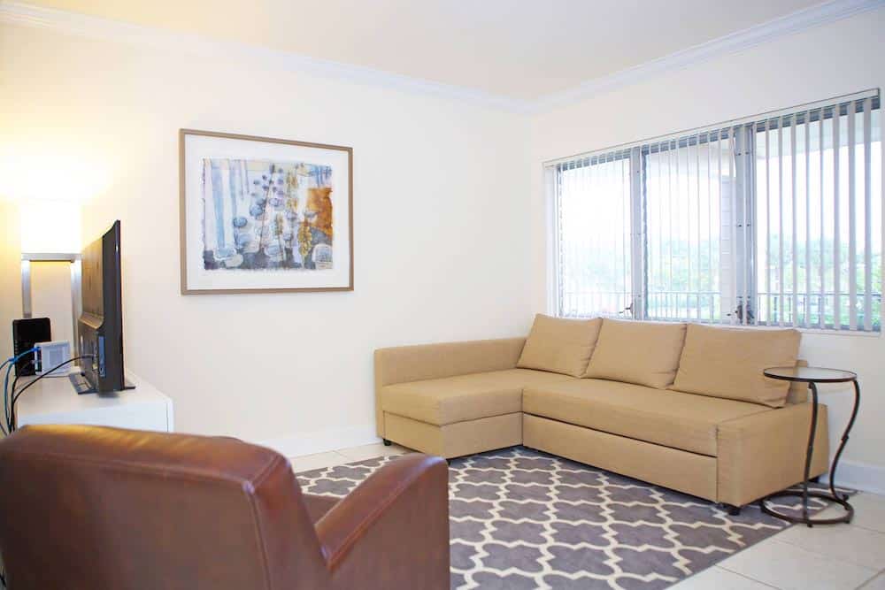 A bright living room featuring a beige sectional sofa, a patterned gray rug reminiscent of Martinique style, paintings on the wall, a TV on a stand, and a brown armchair. Large windows with vertical blinds allow natural light to enter the space. A small round side table sits nearby.