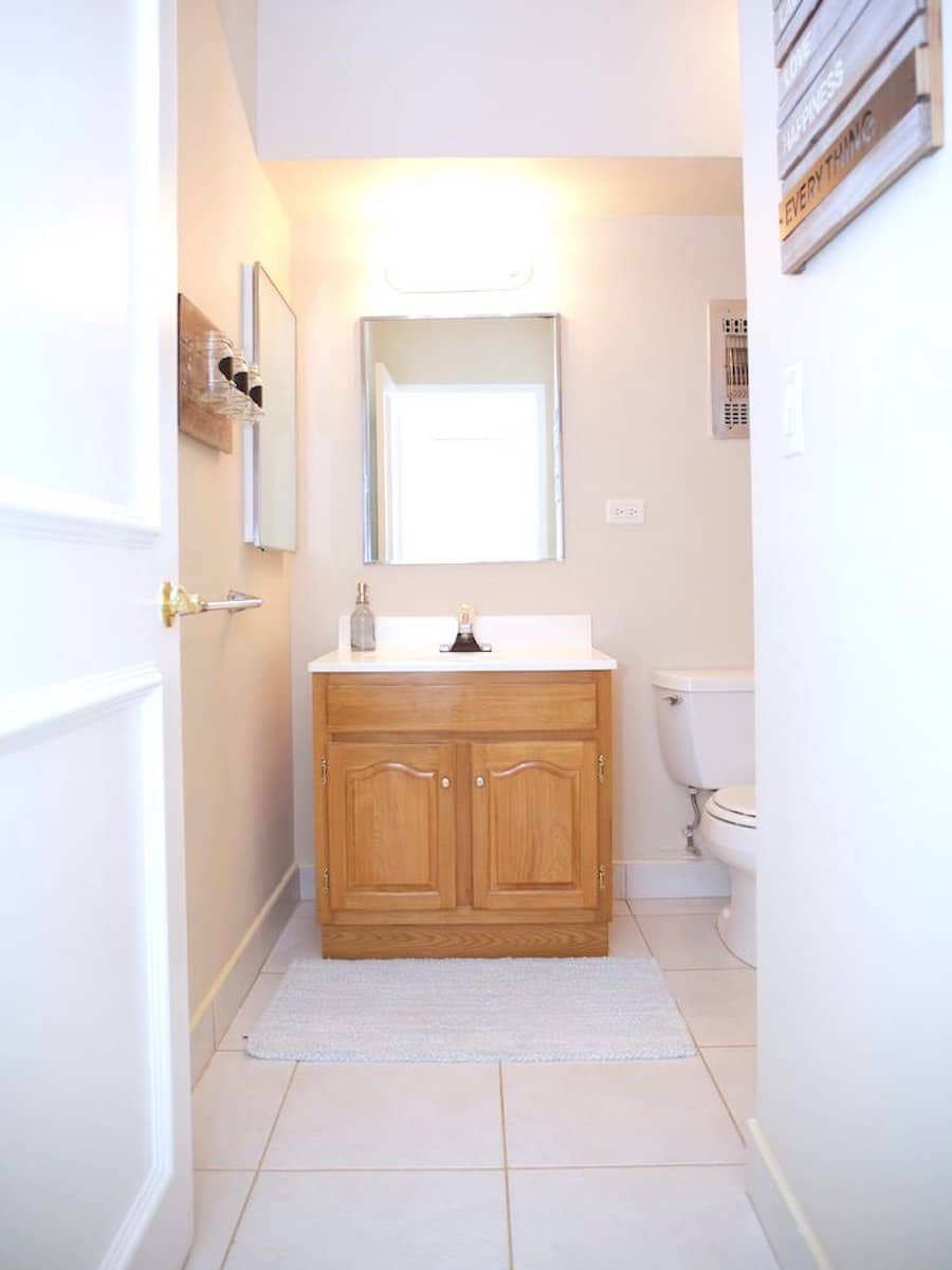 A small bathroom with light beige walls, reminiscent of a serene retreat in Martinique. It features a wooden vanity with a white sink, a rectangular mirror above, and a toilet to the right. Bright overhead lighting and a woven mat add warmth in front of the vanity.