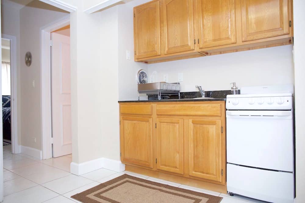 A compact Martinique-inspired kitchen with wooden cabinets, a small white stove, and a stainless steel dish rack on the counter. The floor is tiled, and a beige rug is placed in front of the stove. An open doorway leads to an adjacent room.