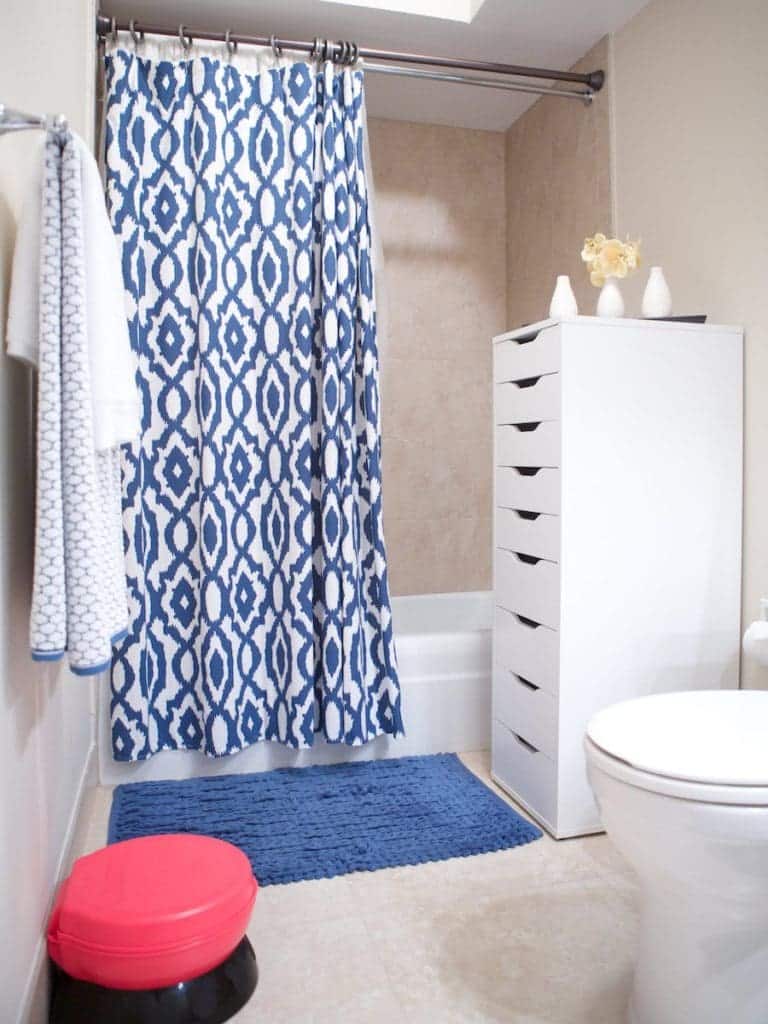 A bathroom reminiscent of Barcelona charm features a blue and white patterned shower curtain, a white cabinet with drawers, a red stool, and a blue bath mat. Elegantly tied together with beige tiles, the space is completed by minimal accessories and a towel hanging on the wall.