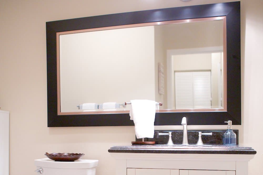 A bathroom features a large rectangular mirror with a black frame above a white sink. The countertop holds a soap dispenser and a folded towel. A toilet is visible on the side, and the background shows an open door and a hallway.