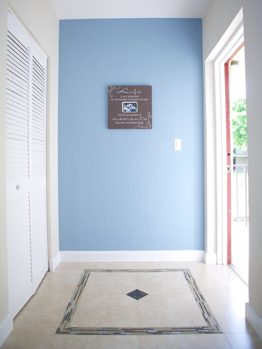 A hallway with a blue accent wall displaying a framed quote reminiscent of Barcelona's artistic flair. The floor features a decorative tile pattern, echoing the city's vibrant style. A white closet door is on the left, while an open door on the right lets in natural light.