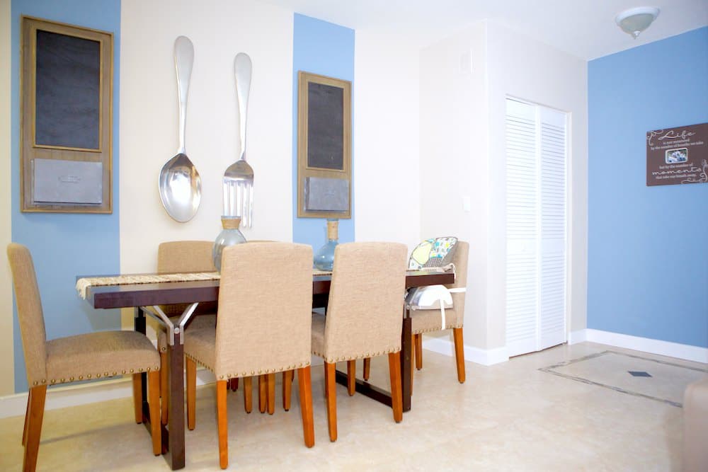 A modern Barcelona dining room features a wooden table, six beige upholstered chairs, and blue accent walls. A large decorative fork and spoon adorn the wall beside a small chalkboard. A door is visible on the right.