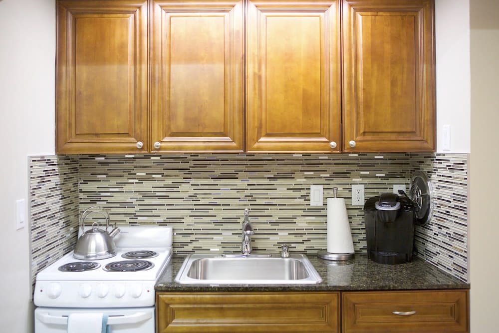 A cozy Barcelona-inspired kitchen with wooden cabinets, a stainless steel sink, and an electric stove hosting a kettle. A coffee maker and paper towel holder rest on the granite countertop, while the backsplash boasts a vibrant multicolored tile design that echoes Barcelona's artistic flair.