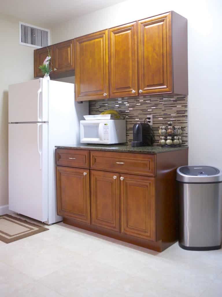 A Barcelona-inspired kitchen corner with a wooden cabinet setup featuring a refrigerator, microwave, and coffee maker. The backsplash is tiled, and there's a stainless steel trash can on the right. The floor is light-colored tile reflecting the city's modern yet traditional charm.