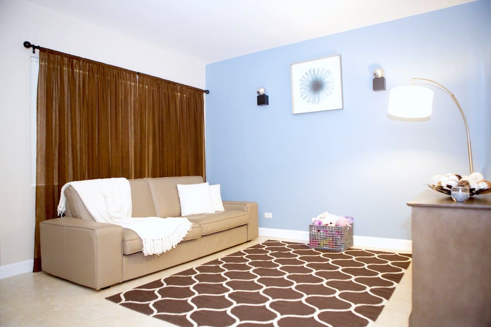 A modern living room with a beige sofa, draped with a white blanket, and two white pillows. It features a light blue accent wall, brown curtains, and a brown and white patterned rug. A floor lamp and a basket of toys are in the corner.