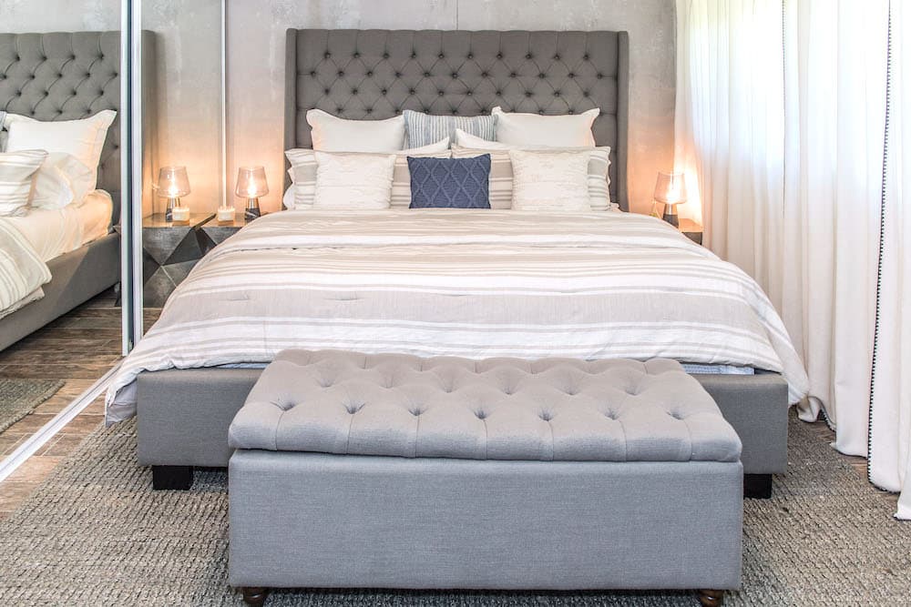 A stylish bedroom featuring a gray upholstered bed with a tufted headboard, adorned with white and gray pillows. A matching gray bench sits at the foot of the bed. The room includes mirrored closets, side lamps, and soft drapes.