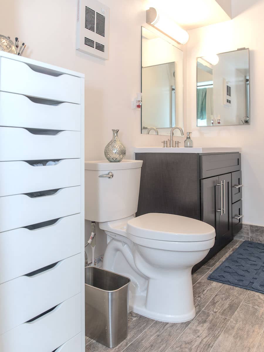 Modern bathroom with a white toilet, a dark vanity with a sink, and a large mirror above. A silver vase sits on the toilet tank. A white drawer unit is on the left, and a small gray rug is on the wooden floor.