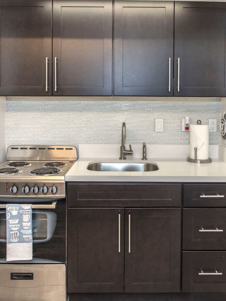 Modern Madison kitchen with dark wooden cabinets, a stainless steel sink, and an electric stove. The white countertop complements the sleek design. A silver faucet is installed over the sink, while a paper towel holder adds practicality to the counter space.