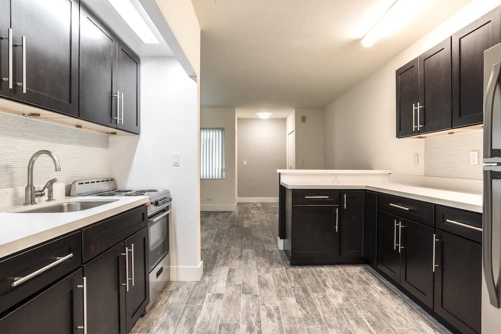 A modern kitchen in Madison, featuring dark wooden cabinets, white countertops, and stainless steel appliances. The floor is adorned with grayish tiles, while a fluorescent light illuminates the space. This stylish room seamlessly extends into a hallway.