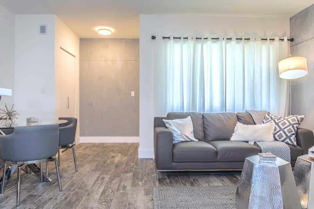 A modern living room with a gray sofa topped with pillows, a floor lamp, and a geometric side table. Sheer curtains cover the window, and a dining area with gray chairs is visible. The room has wooden flooring and neutral colors.