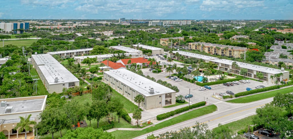 Aerial view of a suburban neighborhood featuring several apartment buildings with white roofs, a central red-roofed structure, a swimming pool, and surrounding greenery. Roads and additional buildings are visible in the background under a partly cloudy sky.