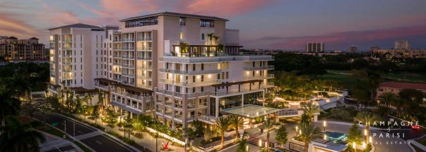 A modern multi-story luxury apartment building at sunset, surrounded by palm trees and a well-lit entrance. The sky displays hues of pink, orange, and blue. Nearby, a pool area is visible, and distant buildings are in the background.