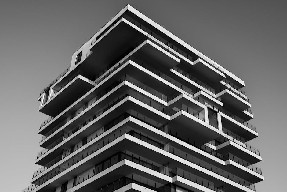 Black and white image of a modern, multi-story building with a unique, staggered design. The structure features overhanging balconies and a geometric facade, set against a clear sky.