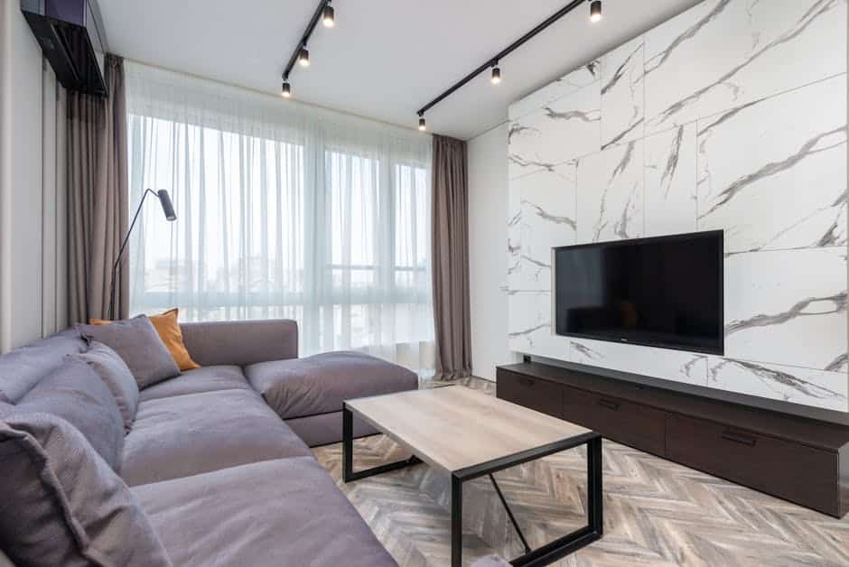 Modern living room with a large gray sectional sofa, marble-patterned feature wall with a mounted flat-screen TV, and a wooden coffee table. Tall curtains cover the window, and track lighting is installed on the ceiling.