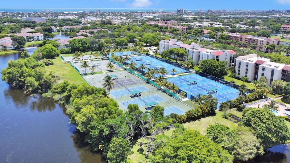 Aerial view of a residential area with several tennis courts surrounded by trees. There are buildings nearby and a body of water. The distant skyline includes more buildings and a glimpse of the ocean under a clear blue sky.