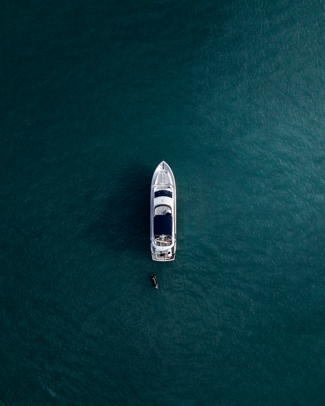 Aerial view of a lone boat anchored on calm, deep blue-green water. A small vessel is positioned next to the boat, casting a shadow. The scene conveys a sense of tranquility and solitude in the vast open water.