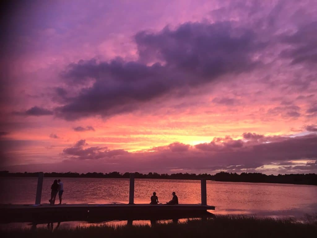 A serene sunset over a lake with dramatic pink and purple clouds. Two couples are silhouetted against the sky, sitting and standing on a dock, enjoying the tranquil view as the sun sets beyond the horizon.