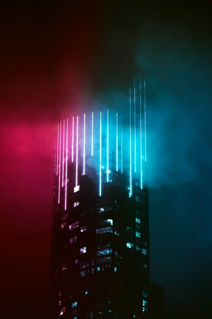 A tall skyscraper at night with glowing vertical lights in pink and blue hues at the top. The lights create a striking contrast with the dark, cloudy sky, giving the scene a futuristic and atmospheric look.